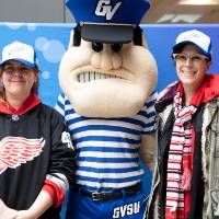 Two alumnae pose, wearing the Red Wings GVSU hat with Louie the Laker at the Detroit Red Wings GVSU Night